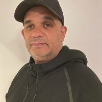 En mann i en svart hettegenser som tilbyr et subtilt smil, selvsikkert poserer mot en nøytral bakgrunn, fremkaller essensen til Rune Ali Zouhar.