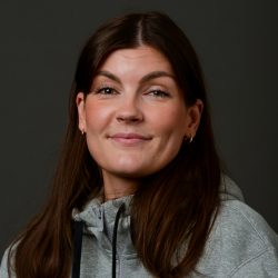 Sara Klæva, en kvinne med et subtilt smil, iført en grå hettegenser, mot mørk bakgrunn.