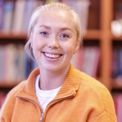 En smilende kvinne med blå øyne og blondt hår, iført en oransje fleecejakke er Veronica Undseth. Hun poserer foran en bokhylle fylt med bøker, og utstråler en munter og kunnskapsrik
