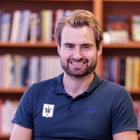 En smilende person iført en blå poloskjorte med logo av Martin Waaler Kaas, poserer foran en uskarp bokhyllebakgrunn.