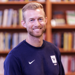 En smilende Vegard Smeby Hammershaug med kort hår, iført en marineblå sportsoverdel med logo, stående foran en uskarp bokhyllebakgrunn.