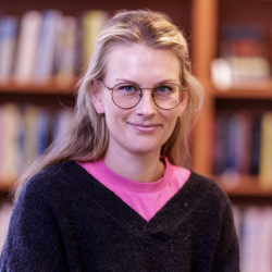 En smilende kvinne med blondt hår og briller, iført en rosa skjorte under en svart genser, står foran en bokhylle. Dette er Kristine Rønning.