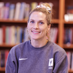 En smilende Thea Therese Næss med håret i hestehale, iført en grå sportsgenser, foran en bokhylle.