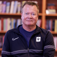 Middelaldrende mann med et selvsikkert blikk, iført en mørk sportsjakke med Pelle Fridell-logo, sittende foran en bokhylle.