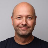 En smilende mann med barbert hode og skjeggstubber, iført svart t-skjorte mot lysegrå bakgrunn, identifiseres som Steffen Uldal.
