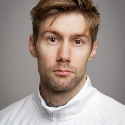 En mann med rufsete hår og et snev av skjeggstubber som ser direkte på kameraet med et nøytralt eller litt perpleks uttrykk.