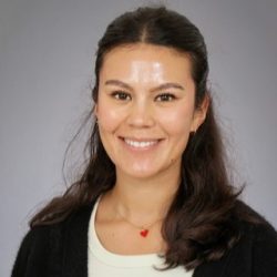 En kvinne med et vennlig smil, minimalt med sminke og et lite rødt hjerteklistremerke på brystet. Håret hennes er mørkt, og hun vises foran en nøytral bakgrunn. Kledd i en
