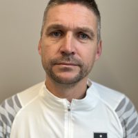 En fokusert person, Tor Trondsen, med kort hårklipp og skygge av skjegg, iført en sporty jakke med glidelås med logo på venstre bryst, ser direkte på kameraet