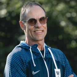 En middelaldrende mann, Bjørn-Morten Moss, med solbriller og blå sportsjakke med hvit logo smiler utendørs.
