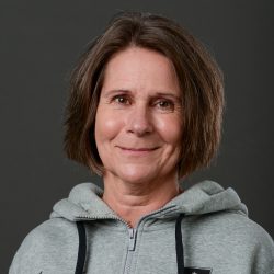 En selvsikker moden kvinne, Mette Smevold Årud, med kort brunt hår, iført en tilfeldig grå hettegenser, smilende forsiktig til kamera mot en mørkegrå bakgrunn.