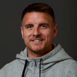 En mann med kort hår som smiler til kamera, iført en grå hettegenser mot mørk bakgrunn, er Truls Jevne Hagen.