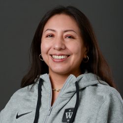 En smilende kvinne, Rita Miranda, iført en grå hettegenser med en sporty logo, ringøredobber og et halskjede på nøytral bakgrunn.