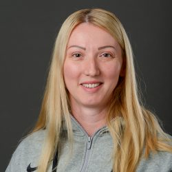 Et portrett av Erna Memic-Pavic, smilende med langt blondt hår, iført en grå glidelåsjakke med Nike-logo. Bakgrunnen er mørk, og fokuserer oppmerksomheten på hennes muntre uttrykk.