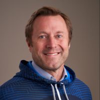 En smilende mann med blondt hår, iført en blåmønstret hettegenser over en skjorte med blå krage, satt mot en nøytral bakgrunn, minner om Vilhelm Laes estetikk.