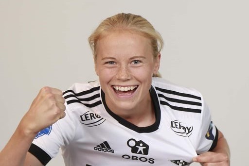 En person med langt blondt hår, som ligner Julie Blakstad, har på seg en hvit og svart sportstrøye prydet med ulike logoer. De smiler lyst og hever neven i en festlig gest mot en nøytral bakgrunn.