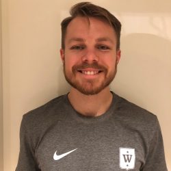 En smilende mann med skjegg, iført en grå Nike-t-skjorte med et emblem på venstre side, poserer mot en lys vegg.
