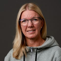 Et portrett av Heidi Høiby, en moden kvinne med blondt hår iført briller og en grå hettegenser, med et subtilt smil, mot en mørk bakgrunn.