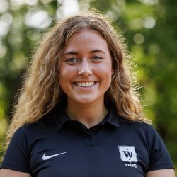 En smilende kvinne med krøllete hår, iført en svart poloskjorte med Kristine Godager-logoen på venstre bryst, stående utendørs med trær i bakgrunnen.
