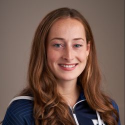 Et portrett av Fredrikke Lovise Hylen, en smilende kvinne med langt, krøllete rødbrunt hår, iført en marineblå atletisk jakke med glidelås og et subtilt mønster, satt opp mot