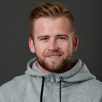 Selvsikker mann med et vennlig smil, pent skjegg og frisyre, uformelt kledd i en grå hettegenser, mot en mørk bakgrunn. Dette er Marius Fauskrud.