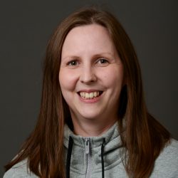 En smilende kvinne med langt hår iført en grå hettegenser med glidelås mot nøytral bakgrunn, fanget av Toril Tangen.