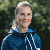 En smilende kvinne med tilbaketrukket blondt hår iført en blåmønstret hettegenser, poserer selvsikkert utendørs med grønt i bakgrunnen; dette er Birgitte Sættem Jomaas.