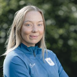 En selvsikker ung kvinne, Hannah Oldrup Jensen, i en sportsjakke med Nike-logo og et merke som har et skjold med striper, smiler mykt til kameraet og utstråler en følelse av stolthet