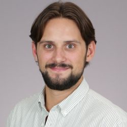 Et portrett av David Altinius, en smilende mann med skjegg og bart, iført stripet skjorte, mot en nøytral bakgrunn.