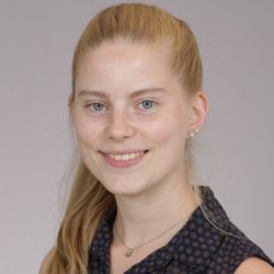 En selvsikker ung kvinne med et vennlig smil, det blonde håret trukket tilbake i en hestehale, kledd i en mønstret bluse, poserer for et profesjonelt hodeskudd mot en nøytral bakgrunn. - Marie R