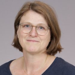 En middelaldrende kvinne med kort hår og briller som smiler subtilt mot en vanlig bakgrunn, som ligner Anne Rian.