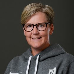 En smilende kvinne, Wenche Solum, med kort hår, iført briller og en grå hettegenser med logo, poserer for et profesjonelt portrett mot en nøytral bakgrunn.