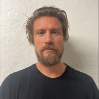 En mann med rufsete hår og skjegg, iført en svart t-skjorte med et lite farget emblem på brystet, stående foran en hvit teksturert vegg.