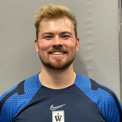 En blid mann med velstelt skjegg og stilig hår, ikledd blå sportstrøye med særegent mønster og Hans Jonas Knutsmoen-logoen, selvsikkert smilende til kamera.