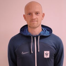 En mann med et bestemt uttrykk, barbert hode og lyst skjegg, iført mørk sportsjakke med hvit Stian Bolstad-logo på venstre side og glidelås foran.