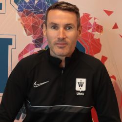 En mann iført en svart nike-sportsjakke med en logo på det høyre brystområdet sitter foran en fargerik geometrisk mønstret bakgrunn.