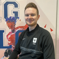 En smilende mann i en svart atletisk jakke med logo, sittende foran et fargerikt grafisk banner med fet skrift.