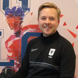 En smilende person som har på seg en svart nike-jakke med en logo (som muligens representerer et idrettslag eller universitet) sitter foran et grafisk bakteppe med geometriske former og mønstre i nyanser av blått og rødt.