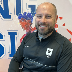 En smilende mann i en svart nike-treningsdress med en logo på brystet sittende foran et levende, geometrisk mønstret bakteppe med bokstavene "ung" synlige.