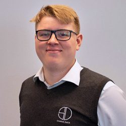 Et profesjonelt hodebilde av en smilende person med briller, iført skjorte og vest med logo, mot en nøytral bakgrunn.