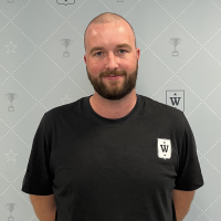 En mann i svart t-skjorte med hvitt emblem smiler lett mens han poserer foran en lys mønstret bakgrunn med stjerne- og trofémotiver.