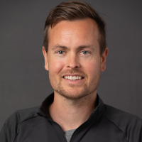 Selvsikker mann med et vennlig smil, iført en uformell svart jakke med glidelås designet av Dag Erik Kokkin, poserer mot en nøytral grå bakgrunn.