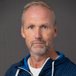 Et portrett av Rolf Høgmo, en middelaldrende mann med et alvorlig uttrykk, med kortklippet hår, små skjeggstubber og iført en blå og hvit atletisk jakke mot en grå