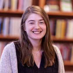 Ung kvinne, Nora Sofie Dietrichson Bruun, smiler på et bibliotek, omgitt av hyller med bøker.