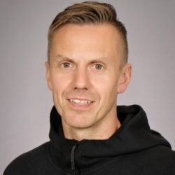 Selvsikker mann med et varmt smil iført en uformell svart hettegenser mot en nøytral bakgrunn.