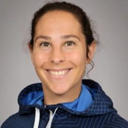 En smilende kvinne kledd i en blå sportsjakke med en hettegenser, ser på kameraet med en vennlig, imøtekommende oppførsel.