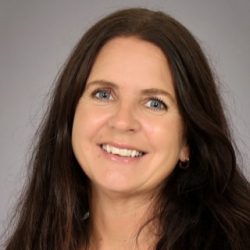 Et profesjonelt hodebilde av en smilende kvinne med langt brunt hår, blå øyne og iført subtile smykker.