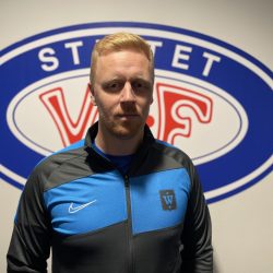 En selvsikker person, Patrick Børresen, poserer foran et idrettslags emblem, iført en Nike-sportsjakke med blå og svart design.
