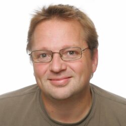 En smilende mann med lyst hår iført briller og en nøytralfarget skjorte mot en vanlig bakgrunn.