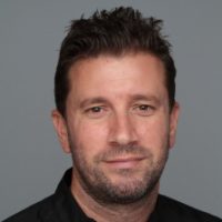 Et portrett av en mann med kort, rufsete hår og lyse skjeggstubber, iført en svart skjorte og tilbyr et subtilt smil med lukket munn mot en grå bakgrunn.