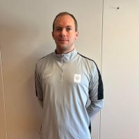En mann, Kristoffer Lie, i en sporty grå Nike-jakke med svarte aksenter står foran en hvit dør og poserer med et nøytralt uttrykk.